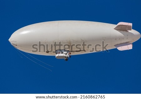 A white blimp, airship, or dirigible flying in blue sky. Close up detail of an unmarked zeppelin like flying vehicle. Flying high above in clear skies.   Royalty-Free Stock Photo #2160862765