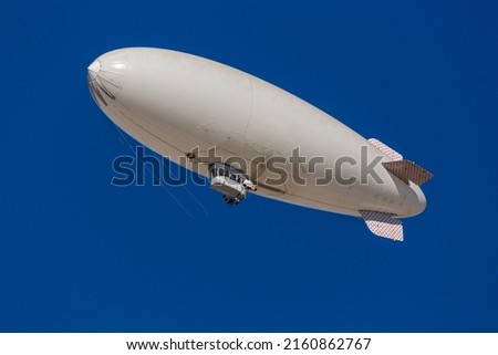 A white blimp, airship, or dirigible flying in blue sky. Close up detail of an unmarked zeppelin like flying vehicle. Flying high above in clear skies.   Royalty-Free Stock Photo #2160862767