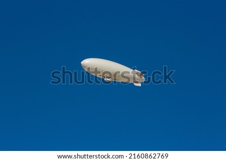 A white blimp, airship, or dirigible flying in blue sky. Close up detail of an unmarked zeppelin like flying vehicle. Flying high above in clear skies.   Royalty-Free Stock Photo #2160862769