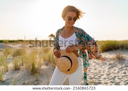 pretty attractive slim smiling woman on sunny beach in summer style fashion trend outfit happy, freedom, wearing white top, jeans and colorful printed tunic boho style chic and straw hat Royalty-Free Stock Photo #2160865723