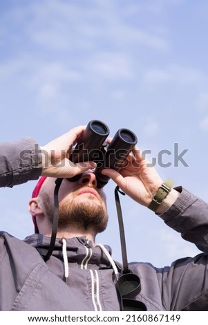 Young man looks up through binoculars against a blue sky, close-up. Birdwatching Royalty-Free Stock Photo #2160867149