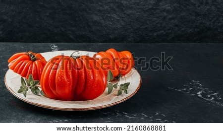 Whole and sliced fresh Raf Coeur De Boeuf, beef tomatoes with oregano on a white plate, grey background, restaurant concept, stock photo