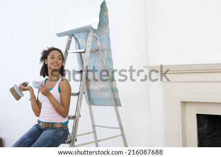 Young woman on ladder with mug taking break from hanging wallpaper, smiling