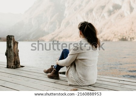 A european woman is sitting on a wooden pier by a lake in the Alps. She is meditating, relaxing and watching towards the hazy mountains. Casual outfit, soft lights. Calm sunset at lake Bohinj.