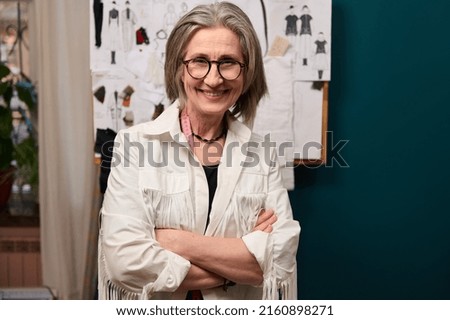 Pleasant stylish elderly European female fashion designer with her arms crossed standing near a wall with fashion sketches and smiles confidently looking at camera