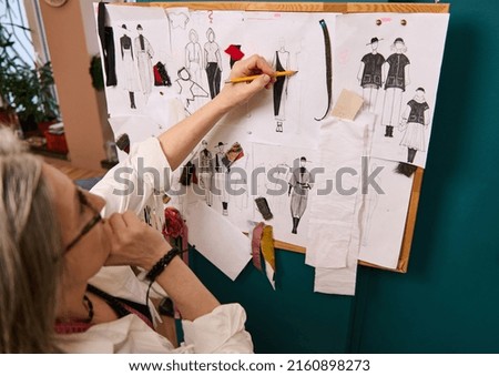 Mature European woman, fashion designer, sketching clothes on the wall with pinned drawings and sketches of a new collection of clothes in the tailoring atelier and fashion design workshop