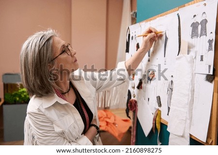 Side portrait of senior woman fashion designer tailor sketching on a paper pinned to the wall with pinned fashion drawings, sketches and templates. Fashion design, tailoring, creative profession