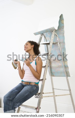 Young woman on ladder with mug taking break from hanging wallpaper, side view