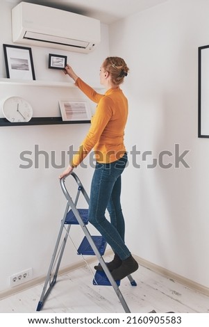 Woman on the ladder hanging pictures and photos on the shelf and wall at home.