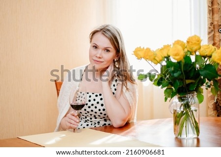 an attractive blonde middle-aged woman with a glass of red wine at a table with a bouquet of yellow roses. home comfort. women's happiness. trainings for single women.