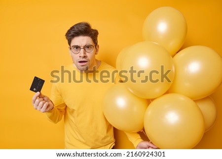 Indoor shot of displeased indignant adult man looks puzzled doesnt know how to make payment online holds credit card and bunch of inflated balloons being cashless isolated over yellow background.