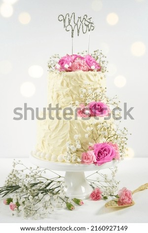 Wedding 2 Tiered Cake with Mr and Mrs topper. Styled with fresh pink roses and gypsophila flowers, against a white background with bokeh party fairy lights. Vertical orientation.