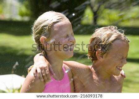 Brother and sister (7-11) arm in arm, smiling with eyes closed, being sprayed with water in garden