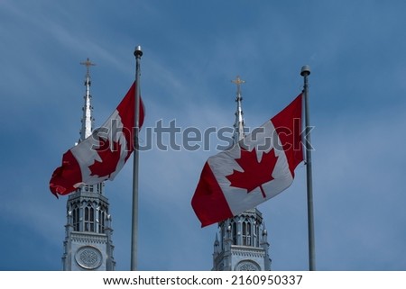 Two Canadian flags waving against blue sky in front of Catholic Church towers.