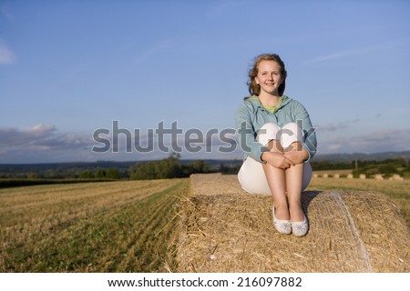 Teenage girl (16-18) on hay bale, knees to chest, smiling, portrait