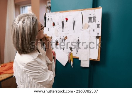 Confident stylish elderly European woman with gray hair thoughtfully looking at a stand with pinned clothes sketches and esquises, standing on her own fashion design workshop. Creating new garment