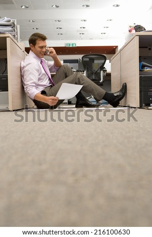 Businessman sitting on floor by piles of paperwork, using telephone, smiling, ground view