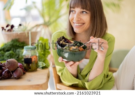 Young cheerful woman eats vegetarian lunch in bowl, sitting by the table full of fresh food ingredients indoors. Healthy lifestyle and wellness concept Royalty-Free Stock Photo #2161023921