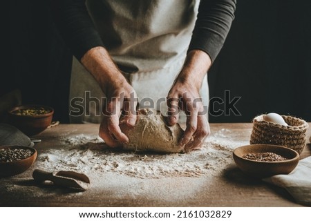 Unrecognizable young man kneading dough on wooden table. Males hands making bread on dark background. Selective focus. Royalty-Free Stock Photo #2161032829