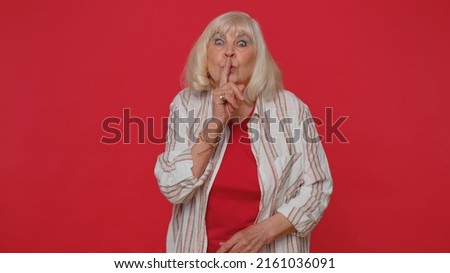Shh be quiet please. Portrait of mature woman 70 years old presses index finger to lips makes silence gesture sign do not tells secret. Elderly senior pretty grandmother on red studio wall background