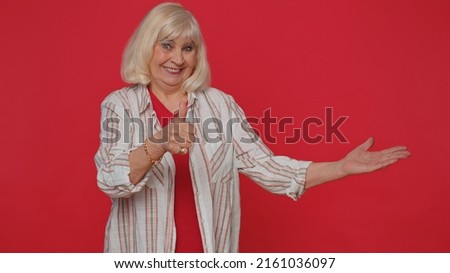 Pretty senior old woman showing thumbs up and pointing empty place, advertising area for commercial text, copy space for goods promotion. Elderly grandmother. Indoors studio shot on red background