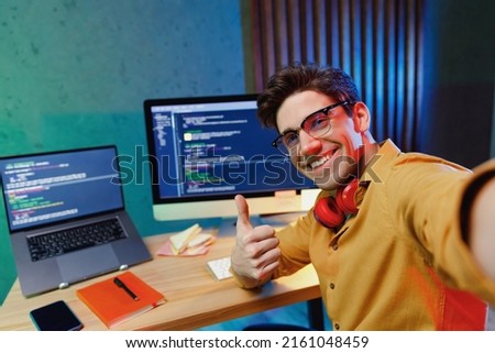 Close up young software site reliability engineer IT programmer man in shirt work at home write code on laptop pc computer do selfie shot pov on mobile phone show thumb up Program development concept