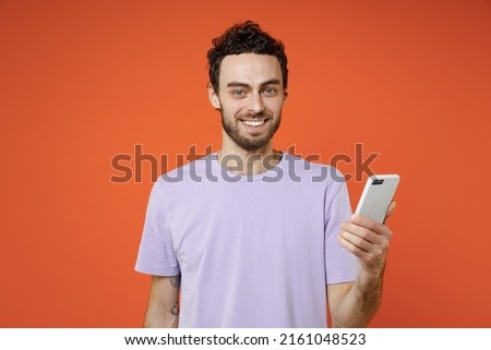 Attractive smiling funny young bearded man 20s in casual violet t-shirt standing using mobile cell phone typing sms message looking camera isolated on bright orange color background studio portrait