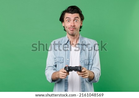 Shocked excited funny cheerful young brunet man 20s wearing casual clothes white t-shirt denim shirt posing standing playing game with joystick isolated on green color wall background studio portrait