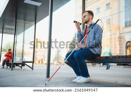 Young blind man with smartphone sitting on bench in park in city, calling Royalty-Free Stock Photo #2161049147