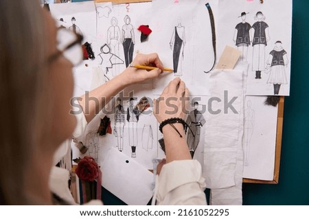 Soft focus on the fashion designer's hands holding a pencil and sketching clothes on paper pinned to the wall with drawings and esquises attached. Creative professional occupation concept