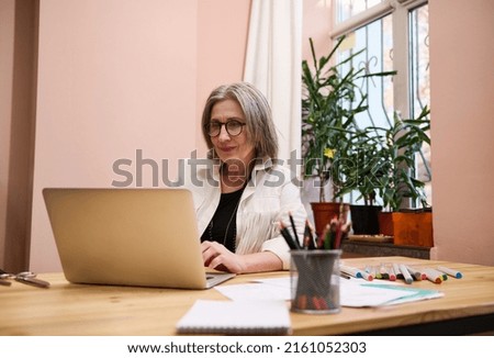 Confident mature European woman, fashion designer seamstress using laptop and designing new clothes for new collection, sitting at a wooden desk in a tailoring atelier