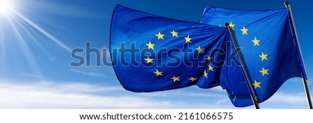 Two European Union flags blowing in the wind on blue sky with clouds and copy space and sunbeams. 