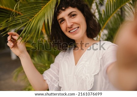 Cute young caucasian girl posing looking at camera taking selfie against backdrop of tropical plants. Brunette wears casual clothes in white. Lifestyle concept