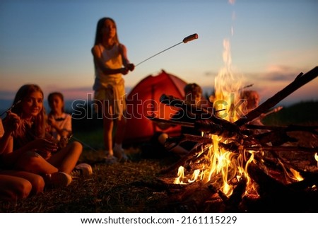 Happy teen girl frying sausage over fire, having fun at campsite at dusk. Side view of group of kids sitting around big campfire, roasting meal at twilight against colorful sky. Concept of camping. Royalty-Free Stock Photo #2161115229