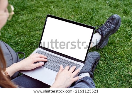 A mockup of a laptop in a girl's lap on the street. A woman works with a laptop in the fresh air in the park, sitting on the lawn. The concept of a remote office. White laptop screen.