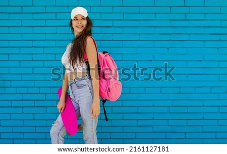 teen sports leisure. young fit brunette girl in jeans with pink skate and backpack stands casual near bllue brick wall background and looks apart with happy smile. lifestyle concept, free space