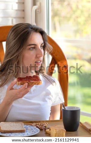 Vertical photo of relaxed young woman sitting having toast and coffee for breakfast as she looks out the window