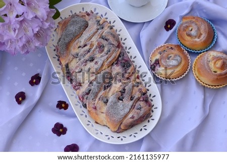 Home baked sweet bread cakes with cheese, berries, and rhubarb. Braided bread roll stuffed with forest fruits, rhubarb, and cheese. Bread buns in baking pans and   rhododendron flower in background.