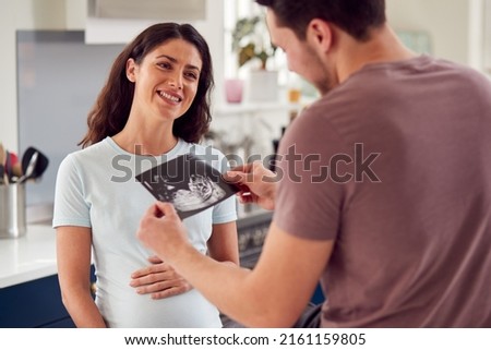 Excited Pregnant Transgender Couple At Home In Kitchen Looking At Ultrasound Scan Of Baby