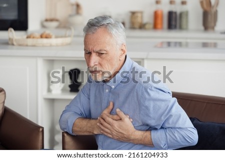Concept of health and cardiovascular problems. Senior man with cardiology disease feeling sharp heart pain, holding hands on chest, sitting on armchair at home. Pensioner need emergency medical care Royalty-Free Stock Photo #2161204933