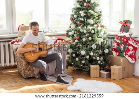 Happy young man is playing guitar. Guy is looking happily and carefree. Male in festive hat alone celebrating Christmas or new year. Christmas tree with garland in background