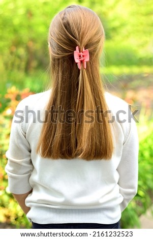 A girl with long blond hair stands backwards against the background of green leaves. Girl in a white sweater.