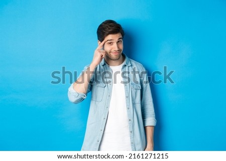 Young thoughtful man pointing at head, asking to think about it, giving a hint, standing in casual clothes over blue background Royalty-Free Stock Photo #2161271215