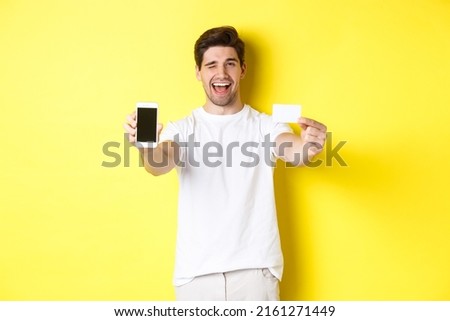 Happy man showing good online offer on mobile phone screen, holding credit card and winking, standing over yellow background