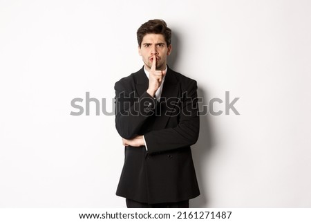 Portrait of angry boss in suit shushing at you, telling to be quiet, showing taboo hush sign and frowning, standing over white background