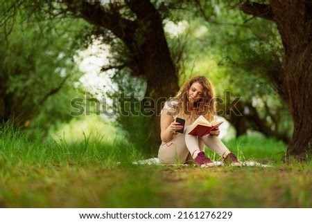 Image of positive caucasian woman lying outdoors in park on a grass and using mobile phone while listening music. she holding book in other hand.