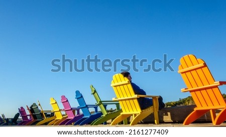 Middle aged man sitting in a row of brightly colored adirondack chairs along waterfront in fall with fall foliage in the distance.