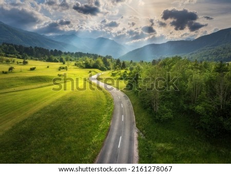 Aerial view of road in green meadows at sunset in summer. Top view from drone of rural road, mountains, forest. Beautiful landscape with roadway, sun rays, trees, hills, green grass, clouds. Slovenia Royalty-Free Stock Photo #2161278067
