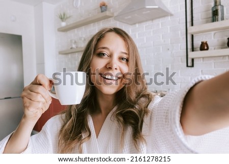 A young beautiful caucasian smiling blonde woman in a white robe with a white cup of coffee or tea takes a selfie in the morning on background of kitchen. Happy girl drinks hot beverage at home Royalty-Free Stock Photo #2161278215