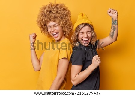 Two positive women friends feel very happy like winners make triumph gesture shake arms celebrate success stand back to each other dressed in casual t shirts isolated over yellow background. Royalty-Free Stock Photo #2161279437
